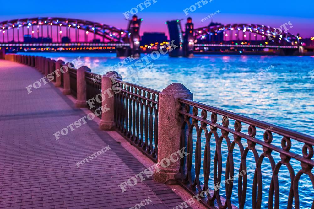 На снимке изображён Большеохтинский мост через реку Неву, подсвеченный вечерним светом. Каменная ограда набережной ведёт взгляд к мосту, который переливается огнями на фоне закатного неба. Этот уголок Санкт-Петербурга символизирует гармонию архитектуры и природы, особенно впечатляющую в вечерние часы.